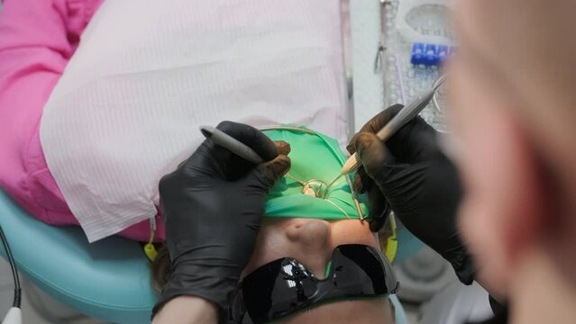 Charming girl in protective glasses and a cheek retractor with a green rubber dam in a dental clinic.