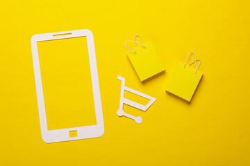 Paper-cut smartphone icon with supermarket trolley and bags on yellow background. Online shopping