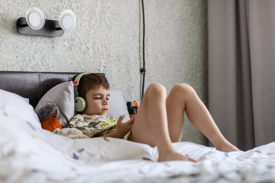 A Boy 6-7 Years Old Brown-eyed Brunette, Listening To Music With Headphones With A Mobile Phone In His Hands, Lying In A Cozy Bed At Home