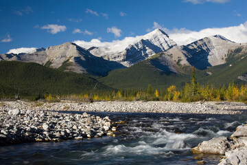 A rushing river with rocky banks and a mountain range with blue sky and clouds in the background in autumn; Bragg creek alberta canada