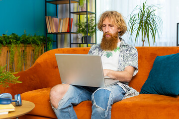 Caucasian redhead man freelancer at home living room apartment and sits down on couch opens laptop. Young bearded guy works on notebook, sends messages, makes online purchases, watching movies