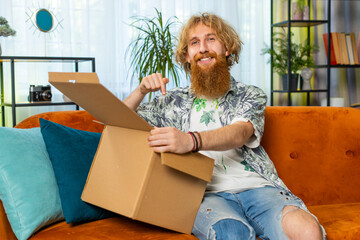 Happy Caucasian redhead man unpacking delivery parcel sitting at home. Smiling satisfied bearded...