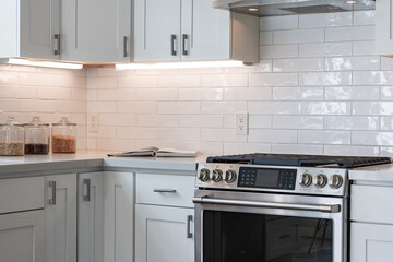 Crisp kitchen details of gas stove, white cabinets, simple white tile backsplash, and marble counter with decorative jars of food.