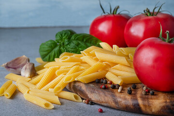 Blurred image of penne pasta, tomatoes, basil on a wooden cutting board on a gray background.