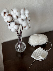 a bouquet of cotton flowers stands in a glass vase on a dark chest of drawers, next to it lies a skein of white wool with a crocheted product.