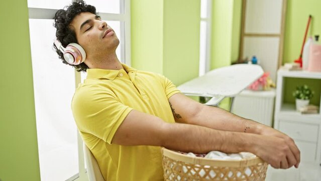 Young latin man holding basket with clothes listening to music relaxed at laundry room