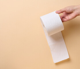A female hand holds a roll of white toilet paper against a beige background