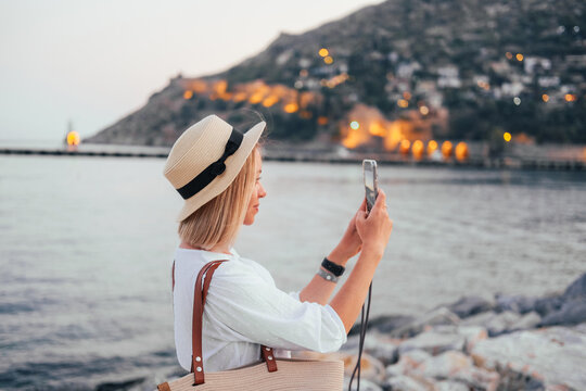 Young woman in a straw hat taking a picture with her smartphone. Sea, evening lights, hills on the background 