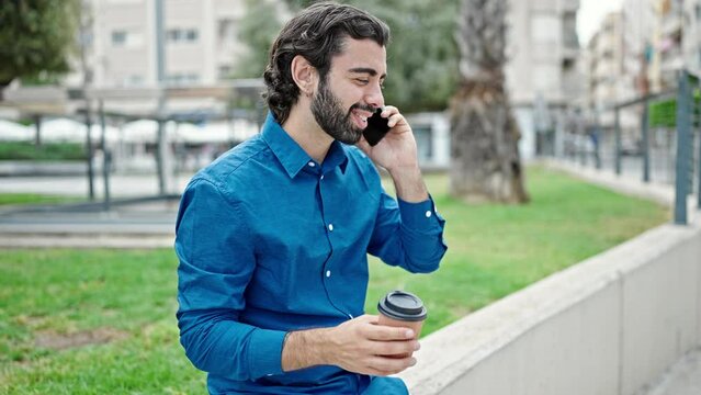 Young hispanic man talking on smartphone drinking coffee at park