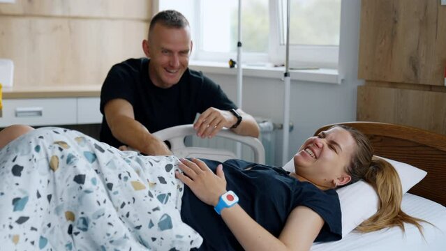 Man in black shirt sits near the bed with his wife. Woman smiles and then feels pain from the contractions. Childbirth concept.