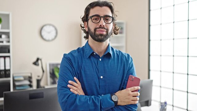 Young hispanic man business worker standing with arms crossed gesture holding smartphone at the office