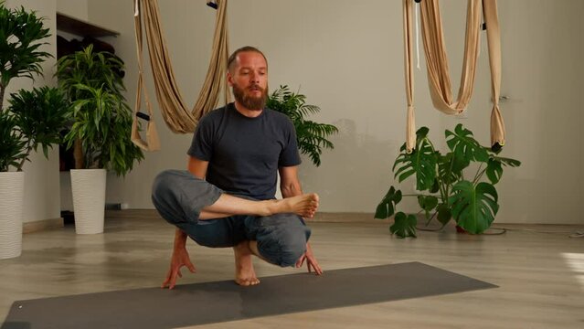 focused adult male yogi with a beard performs a pose exercise from yoga meditates and keeps balance in the hall against the background of flowers