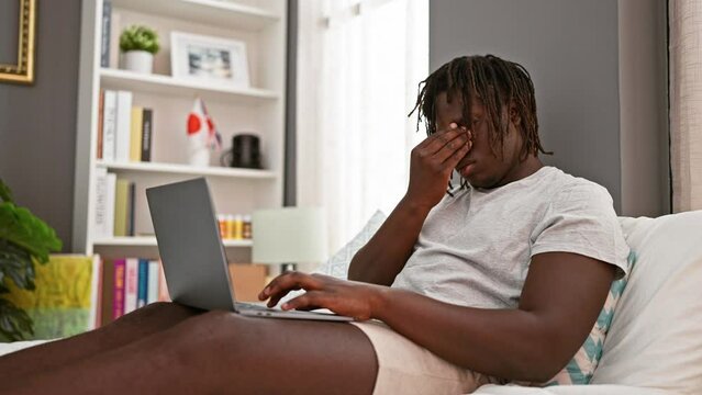 African american man using laptop sitting on bed tired at bedroom