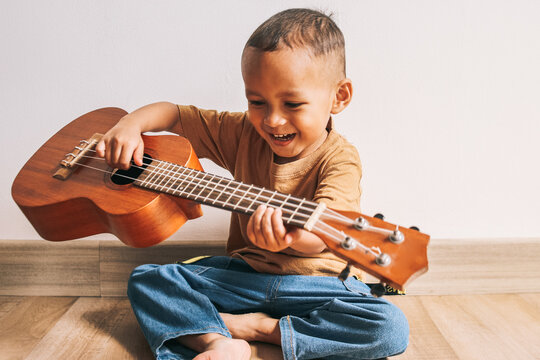Excited Asian little kid sitting on floor and playing guitar ukulele at home