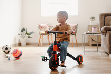 Adorable Asian toddler riding on tricycle at home and looking aside.