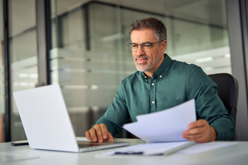 Smiling middle aged executive, mature male hr manager holding documents using laptop looking at pc computer in office at desk, checking financial data in report, doing account paper plan overview.