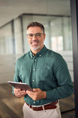 Senior smiling business man holding tablet standing in office. Middle aged businessman manager investor using tab computer, mature male executive looking at camera at work. Vertical portrait.