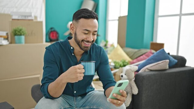 Young hispanic man using smartphone drinking coffee at new home