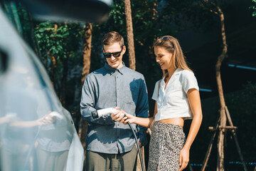 Young couple travel with EV electric car charging in green sustainable city outdoor garden in...