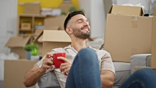 Young hispanic man drinking coffee sitting on floor at new home
