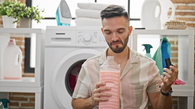 Young hispanic man smelling clean towel and detergent bottle smiling at laundry room