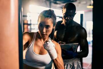 Asian female muay thai boxer with muscular and strong physical body training, punching at boxing bag instructed by her personal trainer or coach in the gym. Martial art and sport concept. Impetus