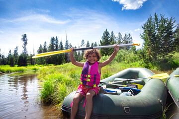 Happy Young diverse boy paddling in an inflatable raft on the river while on a family vacation. Holding his paddle up high in excitement and enjoying the great outdoors - obrazy, fototapety, plakaty