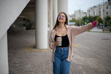 Fototapeta premium a young girl in jeans, a shirt with a mug of coffee and a phone in her hands stands on the street at full height, arms spread wide with joy and smiling