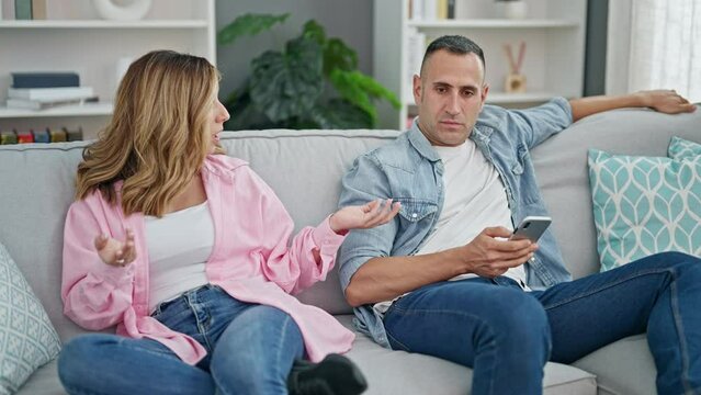 Man and woman couple sitting on sofa using smartphone arguing at home