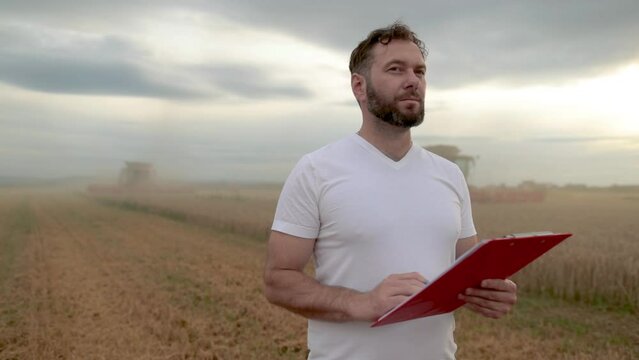A farmer with a tablet for taking notes stands in a field next to combines working in the background. business agriculture concept. Modern agriculture.