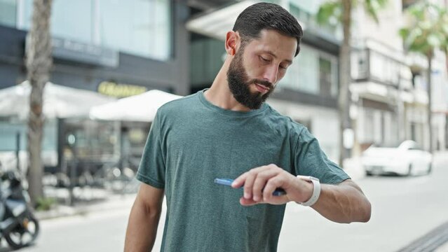 Young hispanic man using earphones looking watch and smartphone at street