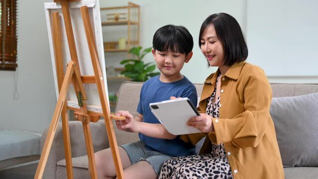Asian Woman assisting young boy in painting on canvas. Happy woman looking at her painting in room