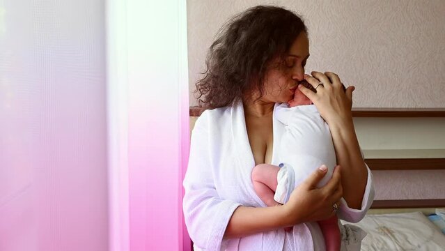 Multi-ethnic middle aged pretty woman, loving caring conscious mother holding her newborn baby burping after feeding milk, standing by window in cozy home bedchamber interior. Maternity leave