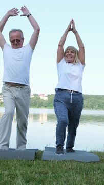 An Elderly Couple Is Doing Sports In The Morning Near The Lake, They Are Doing Stretching Exercises