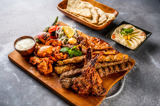 Mix grill with shish tawook, mutton chop, champ, chicken kabab, beef kebab, tikka boti, reshmi and malai, hummus, pita bread, tandoori roti, raita, salad and lemon served in wooden board top view