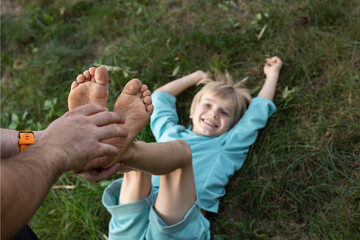 man's hands tickle a child's bare feet lying on the grass. Fun family time together, happy childhood. son's day. The joy of communication, playing around with dad. Top view, selective focus - obrazy, fototapety, plakaty