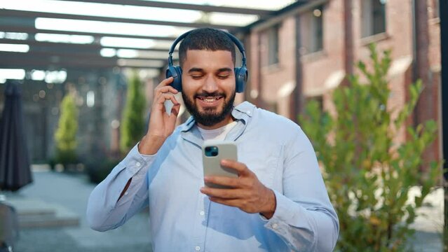 Playful man of indian ethnicity feeling satisfaction from listening to new tracks of favourite rock band. Cheerful person in casual wear and remote headphones hanging out outdoors and enjoying music.