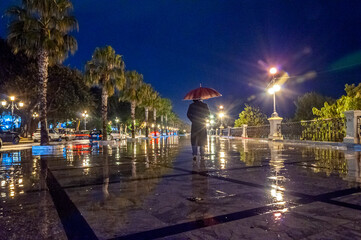 Uomo con cappotto lungo e ombrello passeggia da solo sulla via Marina di Reggio Calabria di notte - obrazy, fototapety, plakaty