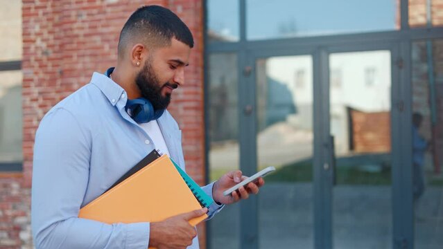 Side view of optimistic indian man strolling on street with notebooks and cellphone in hands and typing message via online application. Carefree male wearing earbuds on neck using wireless technology.