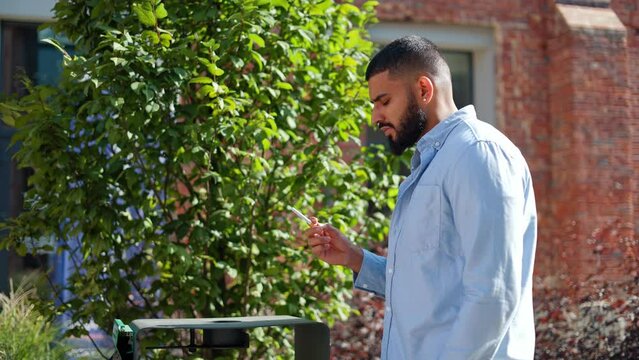 Young indian man in casual outfit walking on street and throwing cigarette into trash can. Bearded gentleman deliberately refusing of smoking. Concept of healthy lifestyle and no smoking campaign.