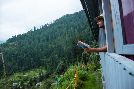young man reading book near window and looking mountain view at countryside homestay in the morning sunrise. SoloTravel, journey, trip and relaxing concept