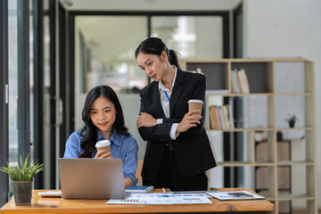 Asian businesswoman holding a mug of hot coffee is discussing work strategy planning. Brainstorm together Find information online on laptop for making reports and summarizing business results.