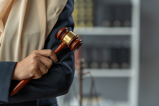 An Asian female Muslim lawyer wearing a hijab stands with her arms crossed and holds the gavel of justice. Confident at work, have law books, study business, laptop Legal scales inside the office.