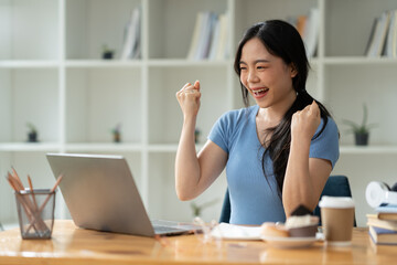 Asian woman sitting at work happily, smiling, delighted with business success feedback data from smartphone and data on laptop. while sitting on a desk in his office at home