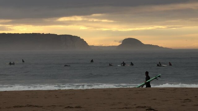 A few people practice surfing in the ocean during the evening against the background of fog breeze, beautiful cliffs and the sunset