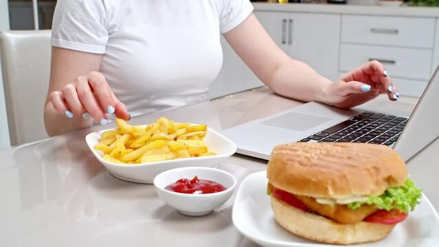 Woman eating unhealthy lunch while studying online courses in the kitchen. Remote work, a freelancer working at home and eating junk food.