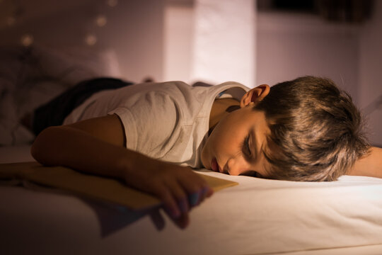 Little cute boy in his children's room in the evening reading a book fell asleep on the bed. Sleepy child hugs a book while lying down sleeping. Active childhood, tired child after a stormy day.