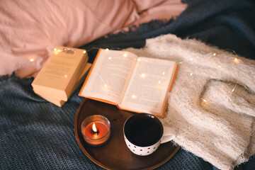 Cup of coffee with open paper book on knit sweater and scented candle on wooden tray close up. Aromatherapy. Cozy home atmosphere. Top view.