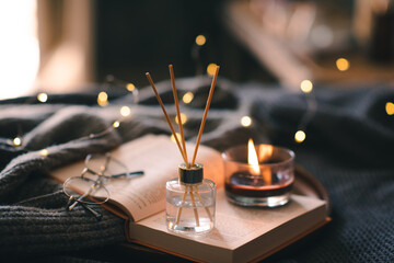 Home liquid fragrance in glass bottle and bamboo sticks with scented candle on wooden tray with open paper book close up over glow lights. Aromatherapy. Cozy home atmosphere.