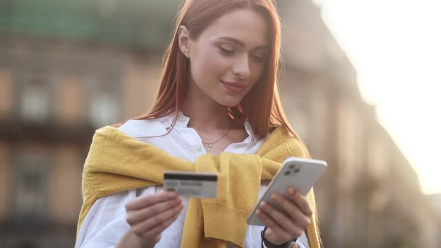 Portrait of charming young woman paying with credit card on smartphone at modern urban city Happy customer doing payments online shopping in internet store and receiving cash back outdoors 
